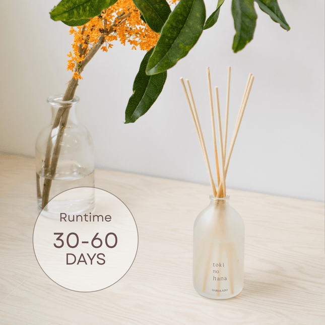 Diffuser bottle with reeds and a vase with flowers on a table, featuring 'toki no hana' branding.