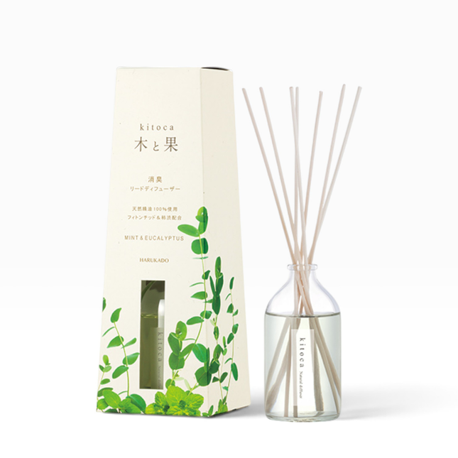 A reed diffuser bottle with light natural reeds next to its cream-colored packaging box decorated with green leaf illustrations. The box is labeled 'kitoca' and 'MINT & EUCALYPTUS'.