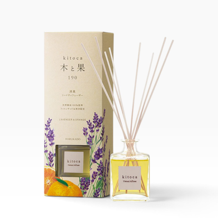 A reed diffuser bottle with pale reeds and yellow liquid stands beside its matching box. The packaging is beige with lavender and orange illustrations, and the bottle label reads kitoca.