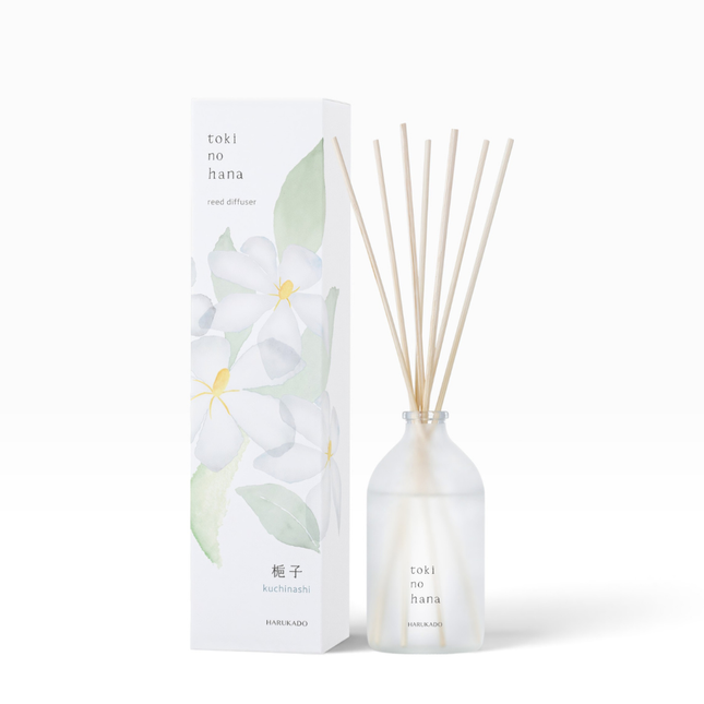 A reed diffuser bottle with natural wood reeds next to its white packaging box decorated with white flowers and green leaves. The box and bottle are labeled 'toki no hana' and 'kuchibiki'.