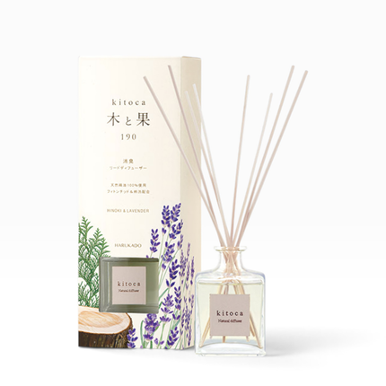 A reed diffuser bottle with light wood reeds stands beside its matching product box. The box is cream-colored with Japanese text, a green glass bottle illustration, and purple lavender drawings.