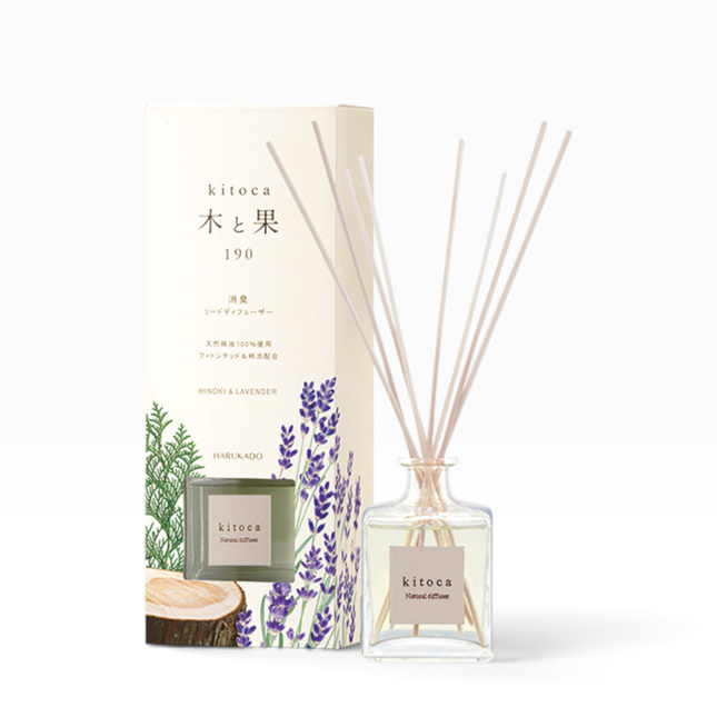 A reed diffuser bottle with light wood reeds stands beside its matching product box. The box is cream-colored with Japanese text, a green glass bottle illustration, and purple lavender drawings.