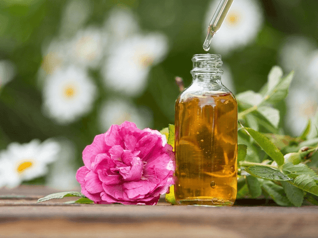 Glass bottle of oil with a dropper and pink flower on a wooden surface with a blurred natural background