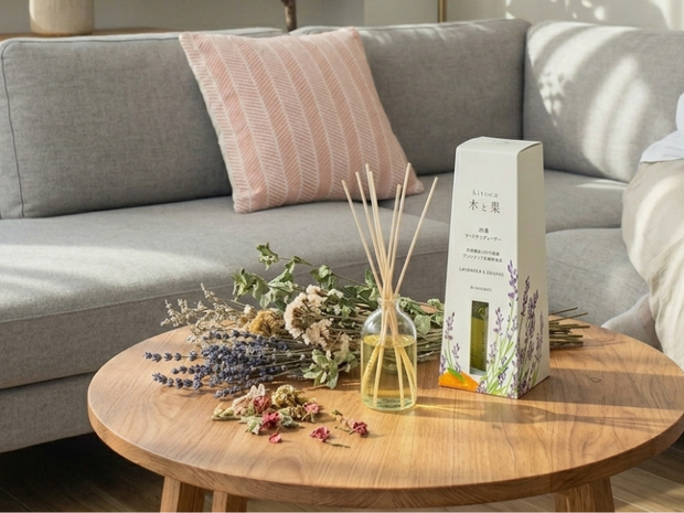 A wooden coffee table with a diffuser, flowers, and a box on it, in front of a gray sofa.